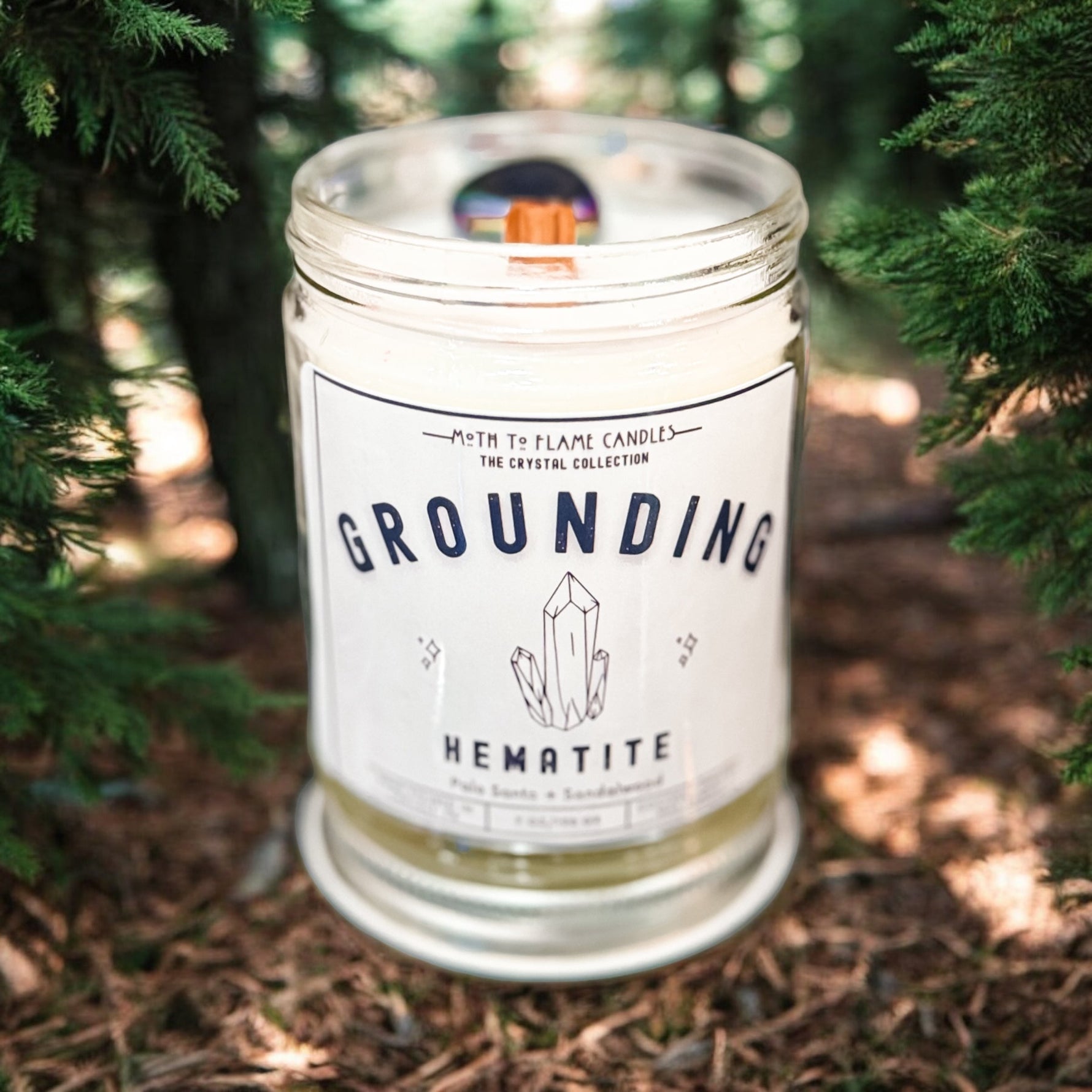 Candle labeled 'Grounding Hematite' in a natural setting with trees.